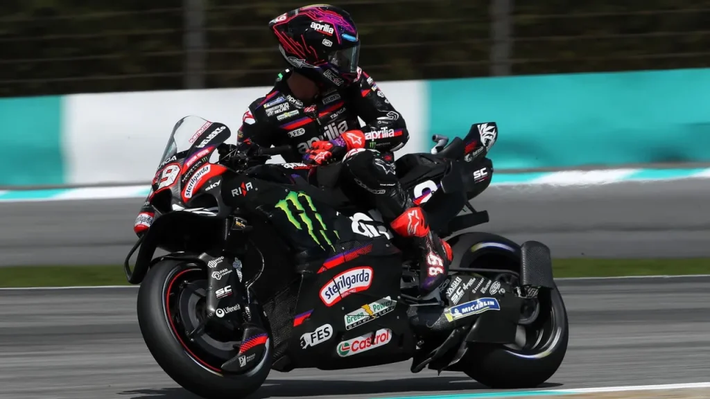 A motorcycle racer in black leathers riding a black Aprilia racing motorcycle on an asphalt track.