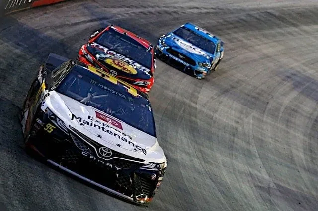 Stock cars race through a turn on a banked oval track.