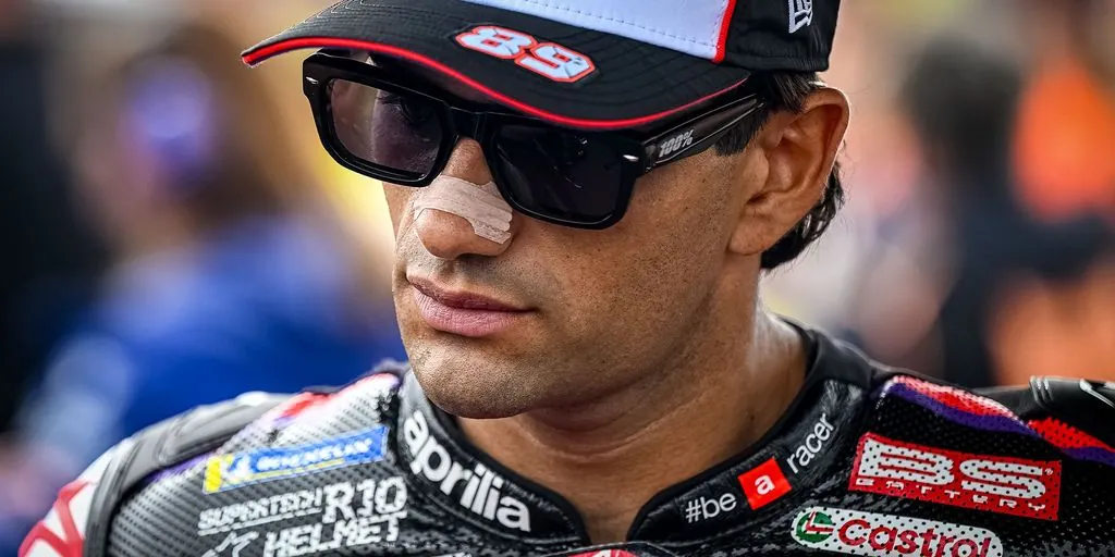A close-up of a motorcycle racer wearing sunglasses, a cap, and racing leathers with a bandage across his nose.