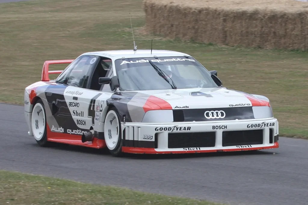 A front-three-quarter view of a white, red, and black #4 Audi 90 Quattro IMSA GTO race car driving on an asphalt course, with grass and straw bales visible in the background.
