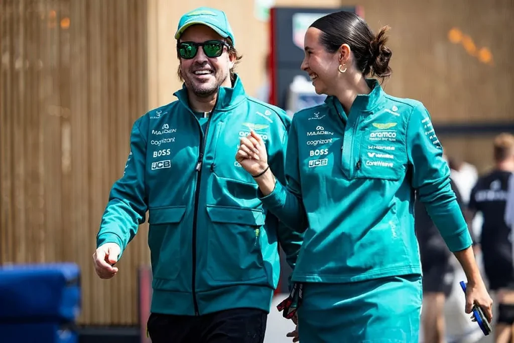 A man wearing a green cap, sunglasses, and team jacket walking alongside a smiling woman in matching team apparel.
