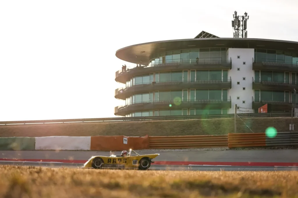 A yellow open-cockpit sports racing car driving on a racetrack in front of a curved multi-story building.