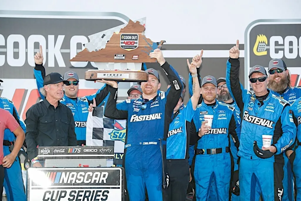 A racing driver in a blue firesuit holds a trophy above his head while celebrating on a podium with team members in matching apparel.