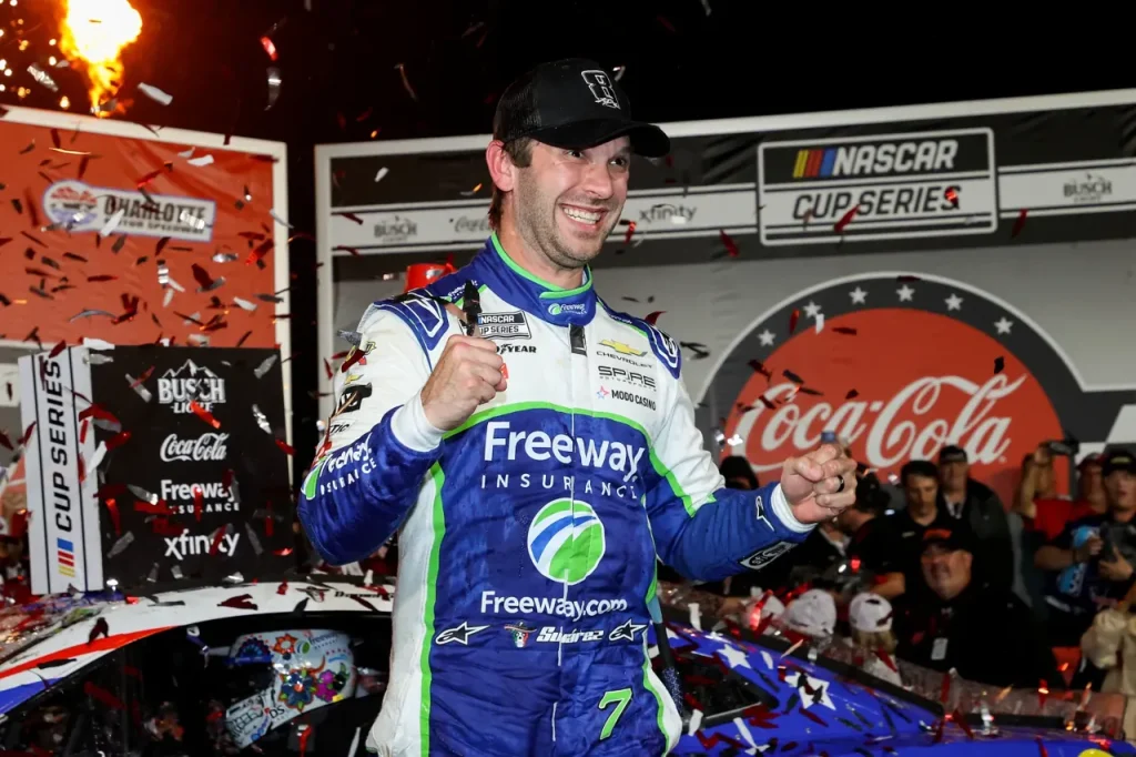 A smiling race car driver in a blue and white racing suit and black cap celebrates with clenched fists amidst falling confetti.