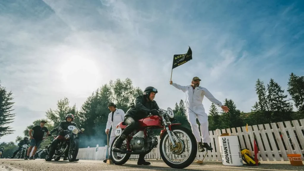 A classic motorcycle and rider at the starting line beside an official in a white jumpsuit jumping into the air while waving an Auerberg Klassik flag.