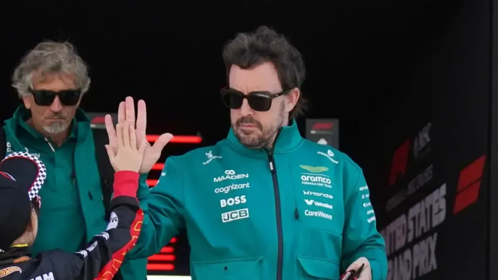 Fernando Alonso wearing sunglasses and a green Aston Martin team jacket high-fiving a person.