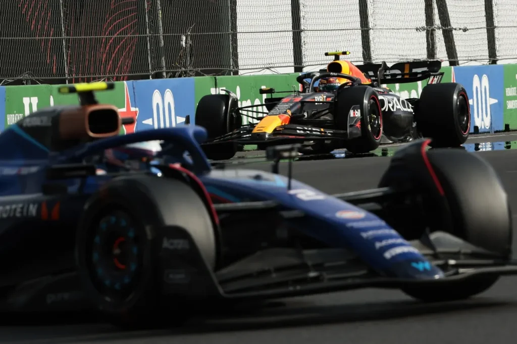 A Formula 1 car on a racetrack ahead of another Formula 1 car alongside trackside barriers.