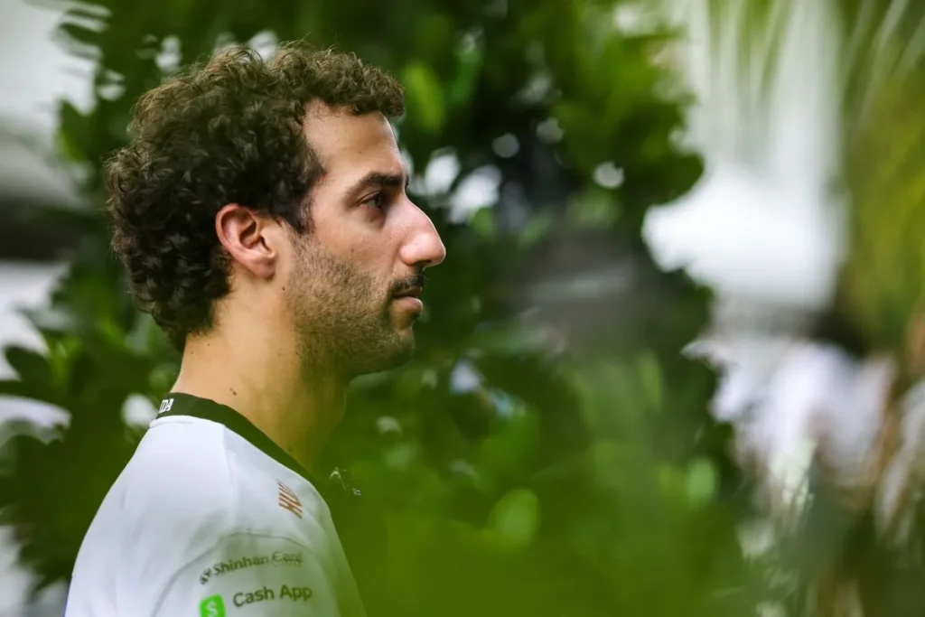 Side profile of a man with curly brown hair and facial stubble wearing a white collared shirt with sponsor logos in front of green foliage.