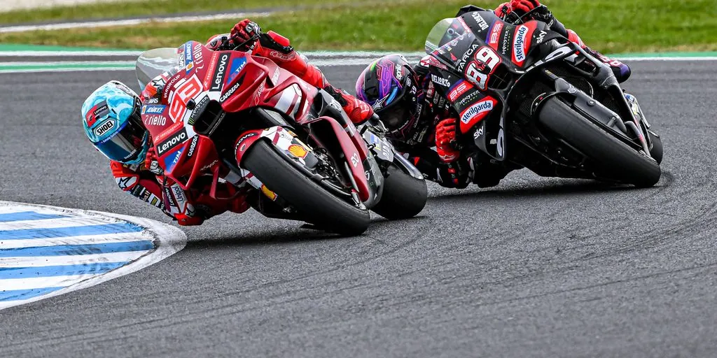 Two motorcycle racers leaning sharply through a turn on an asphalt racetrack, with a red motorcycle leading a black motorcycle.