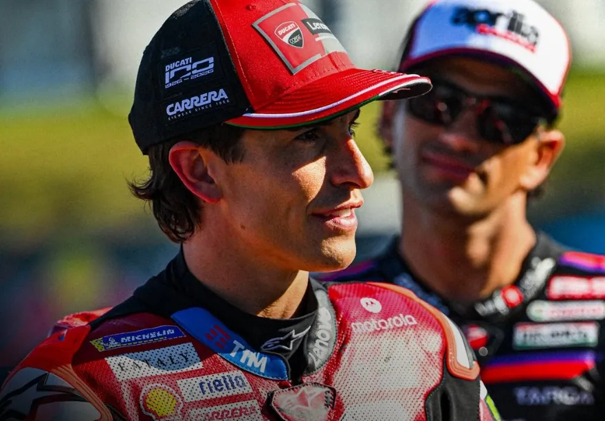 A motorcycle racer in a red cap and racing leathers in the foreground, with another person wearing a cap and sunglasses in the background.