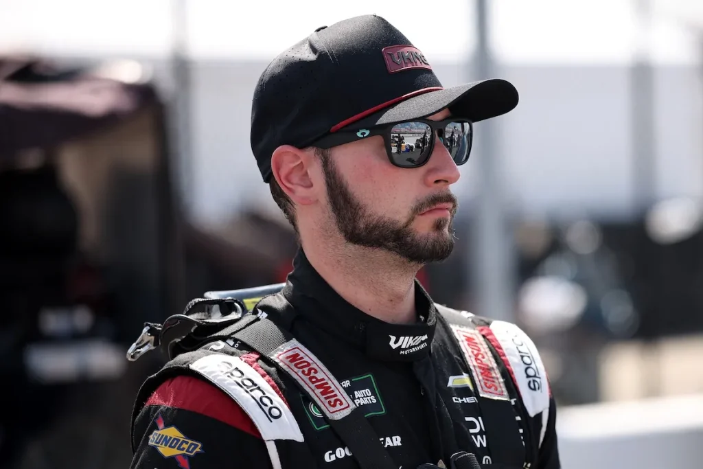 A bearded racing driver wearing sunglasses, a black baseball cap, and a black and red racing suit with Viking Motorsports branding on the collar.