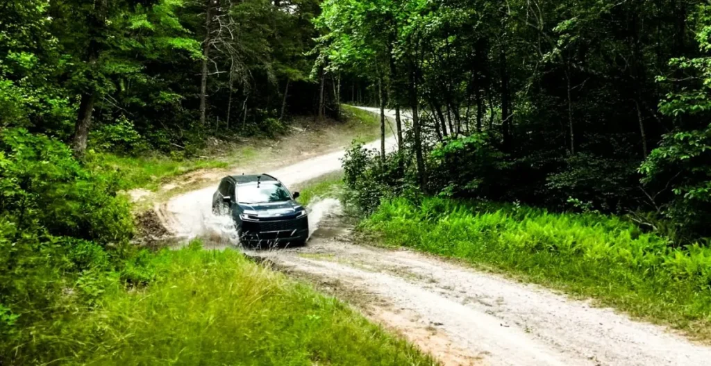 A dark SUV drives through a water splash on a dirt road winding through a dense forest.