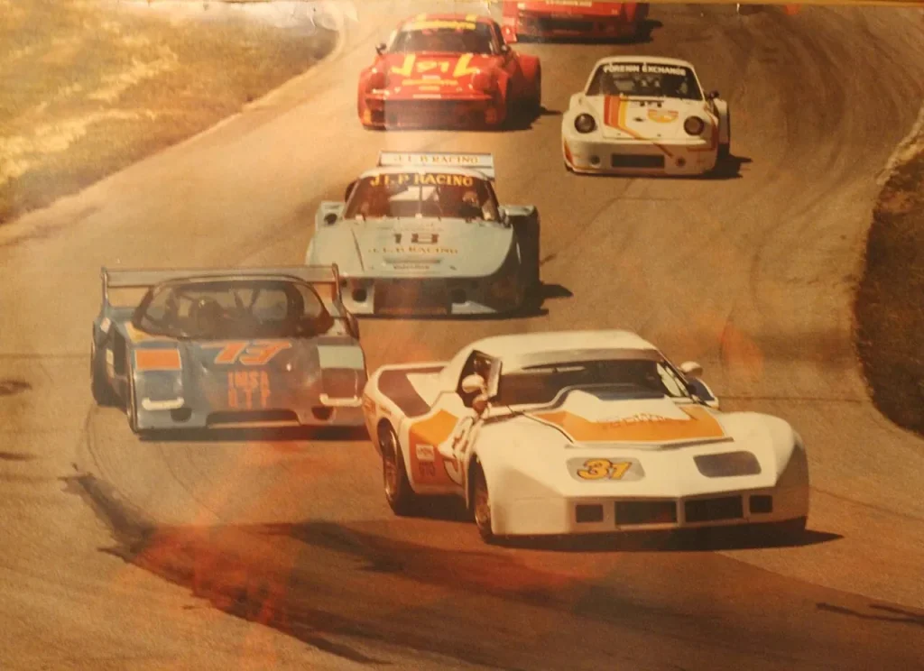 A group of IMSA sports cars navigating a turn on an asphalt circuit, prominently featuring a white and orange Chevrolet Corvette (No. 31) with GTO markings alongside a light blue GTP prototype (No. 73) displaying 'IMSA GTP' lettering on the nose, trailed by a light blue J.L.P. Racing.