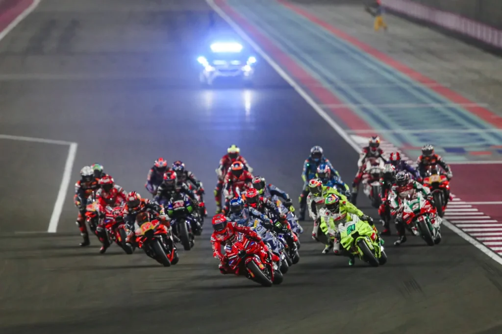A pack of Grand Prix motorcycle riders racing on a circuit at night under floodlights.