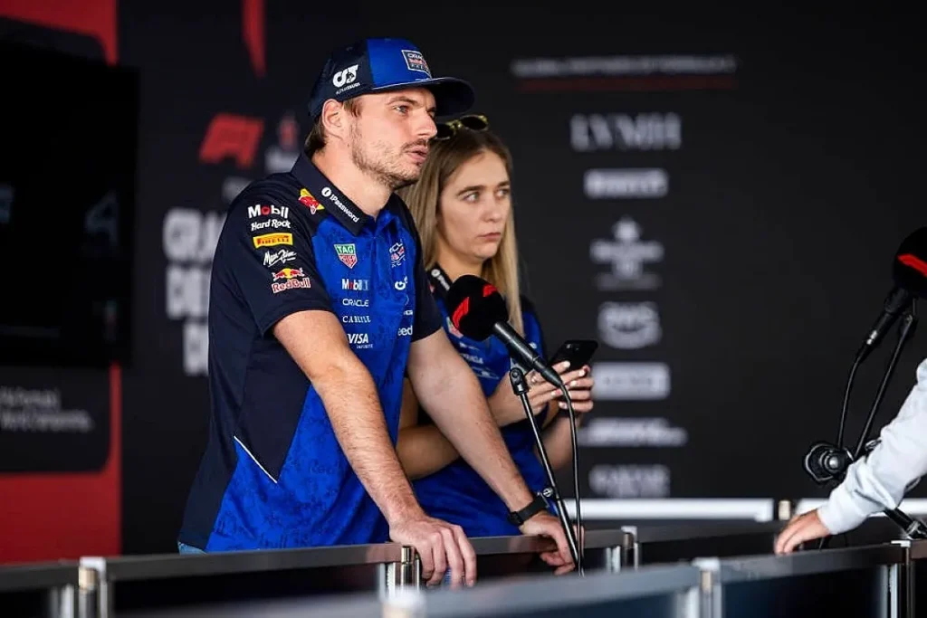 A man in a blue racing team cap and polo shirt leans on a media barrier facing microphones, with a woman standing behind him.