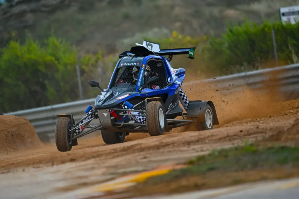 A blue and black CrossCar kicks up dust while racing on a dirt track section.