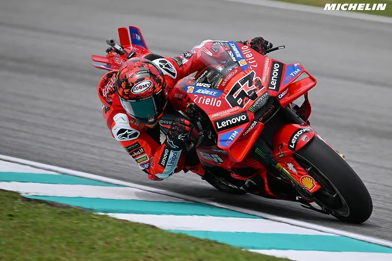A motorcycle racer in a red suit and helmet leaning deeply into a turn on a red racing motorcycle on a paved circuit.