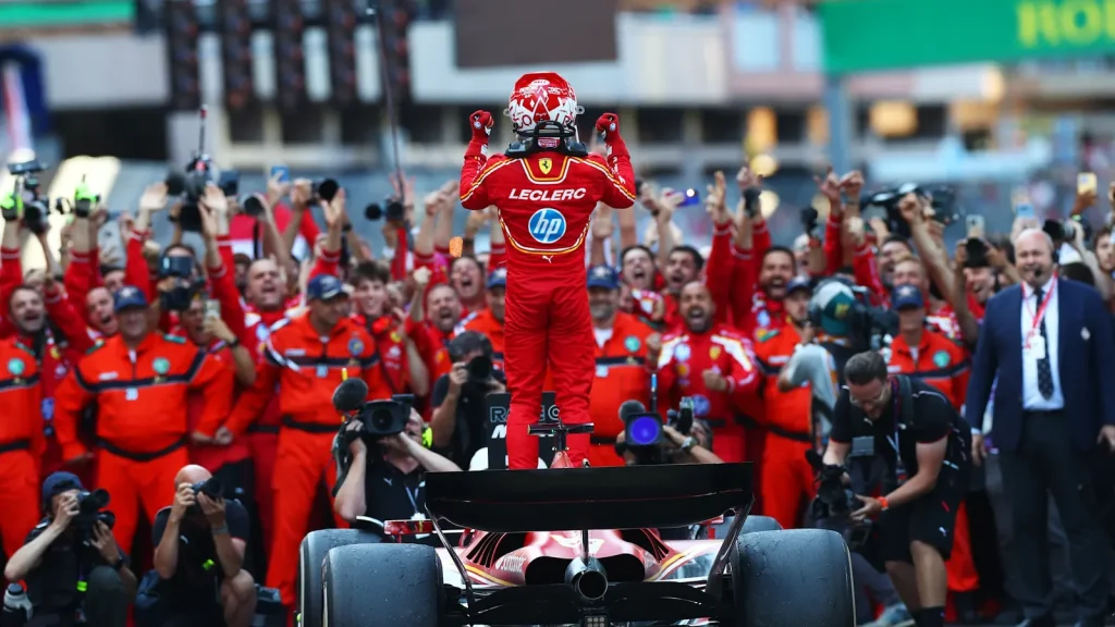 Charles Leclerc stands on a Ferrari with both arms raised while team personnel celebrate and photographers surround the car.