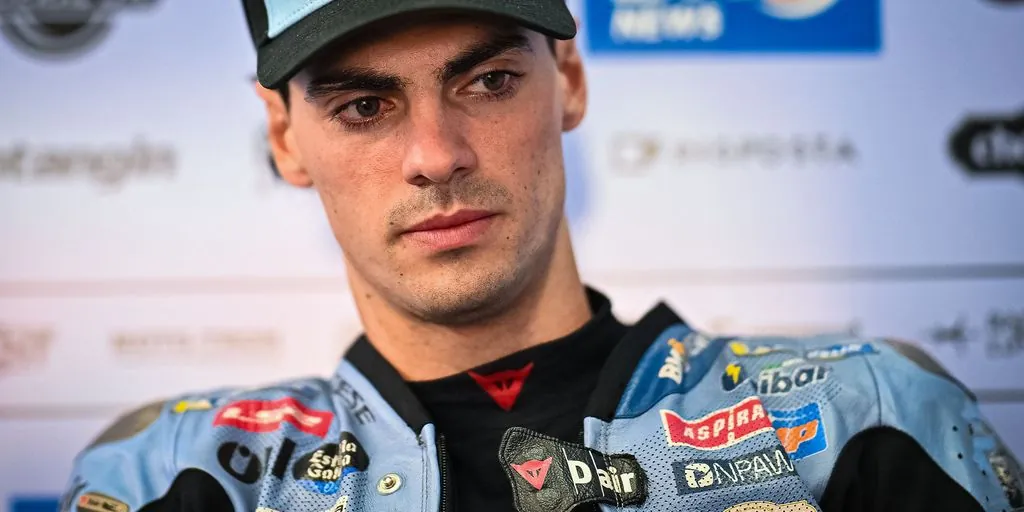 A motorcycle racer wearing racing leathers and a cap looks forward.