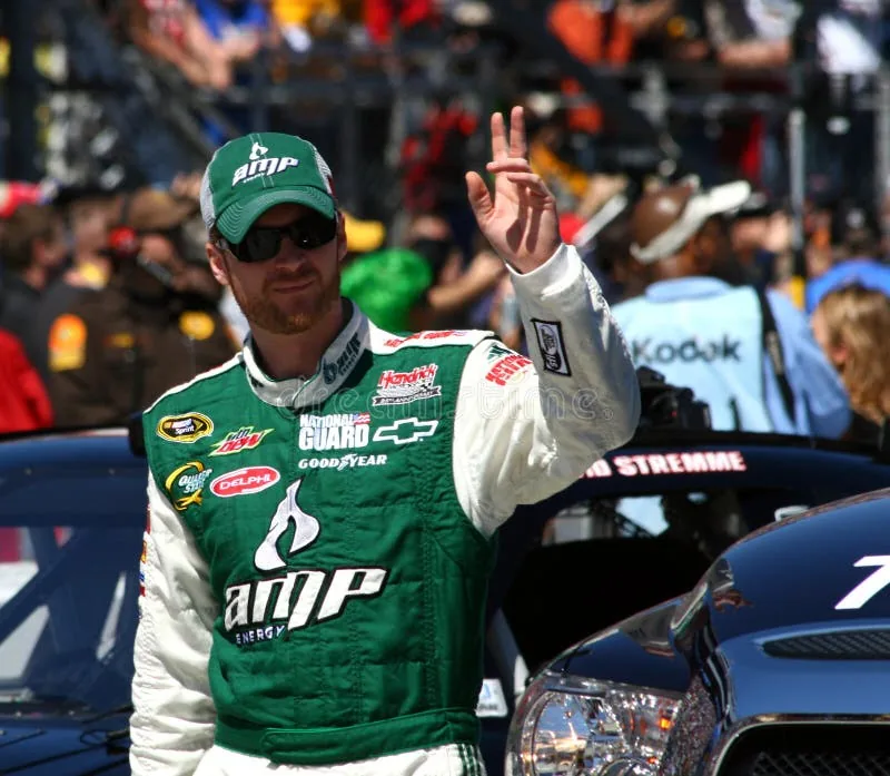 Driver wearing sunglasses, a green cap and a green-and-white AMP racing suit waves beside stock cars, with a crowd behind him.