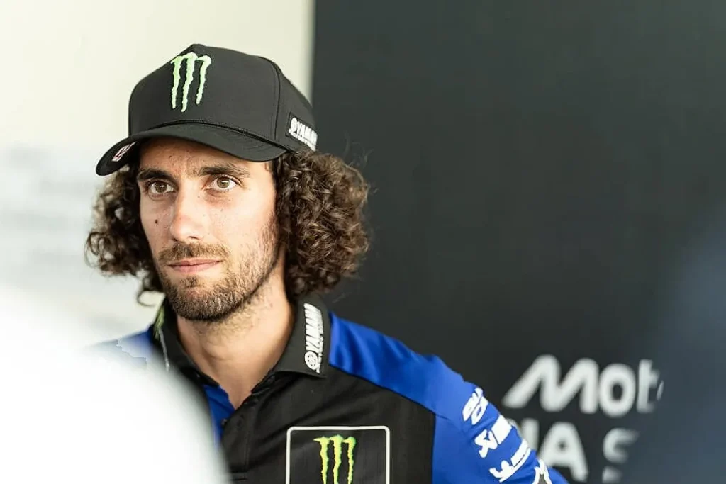 A man with curly dark hair and a beard wearing a black cap and a blue and black collared team shirt.