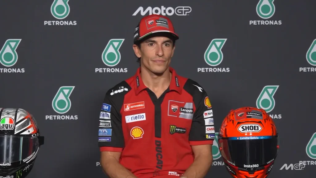 Marc Márquez wearing a red Ducati team shirt and cap seated at a press conference table with racing helmets in front of a MotoGP and Petronas backdrop.