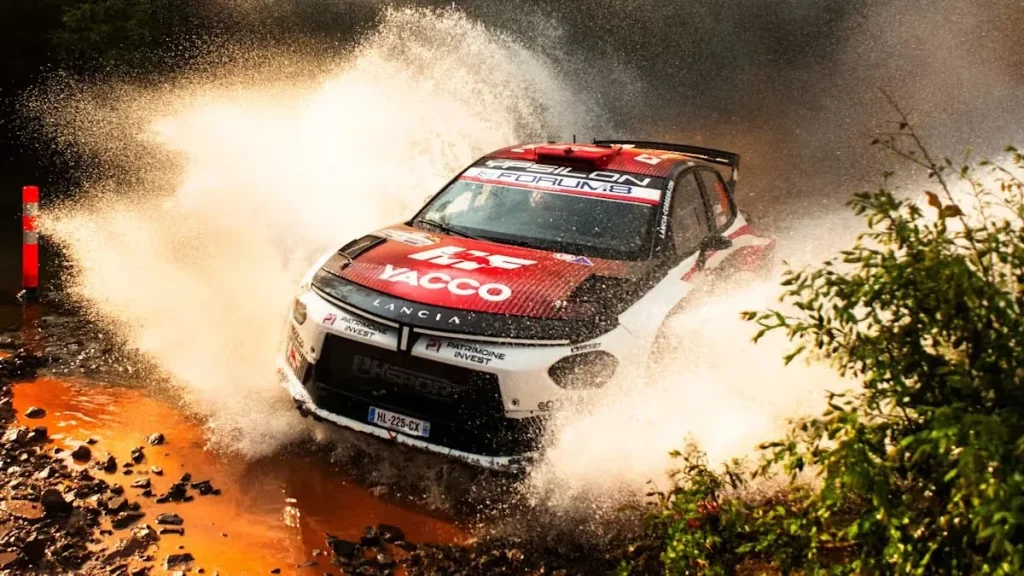 A red and white Lancia Ypsilon rally car driving through a water splash.