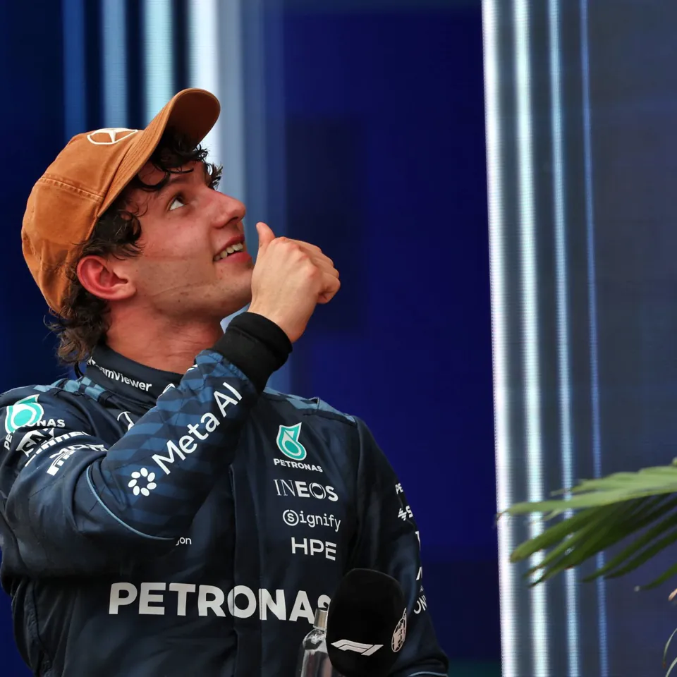 A person wearing a Mercedes racing suit and a tan cap looks upward while gesturing with a thumb.