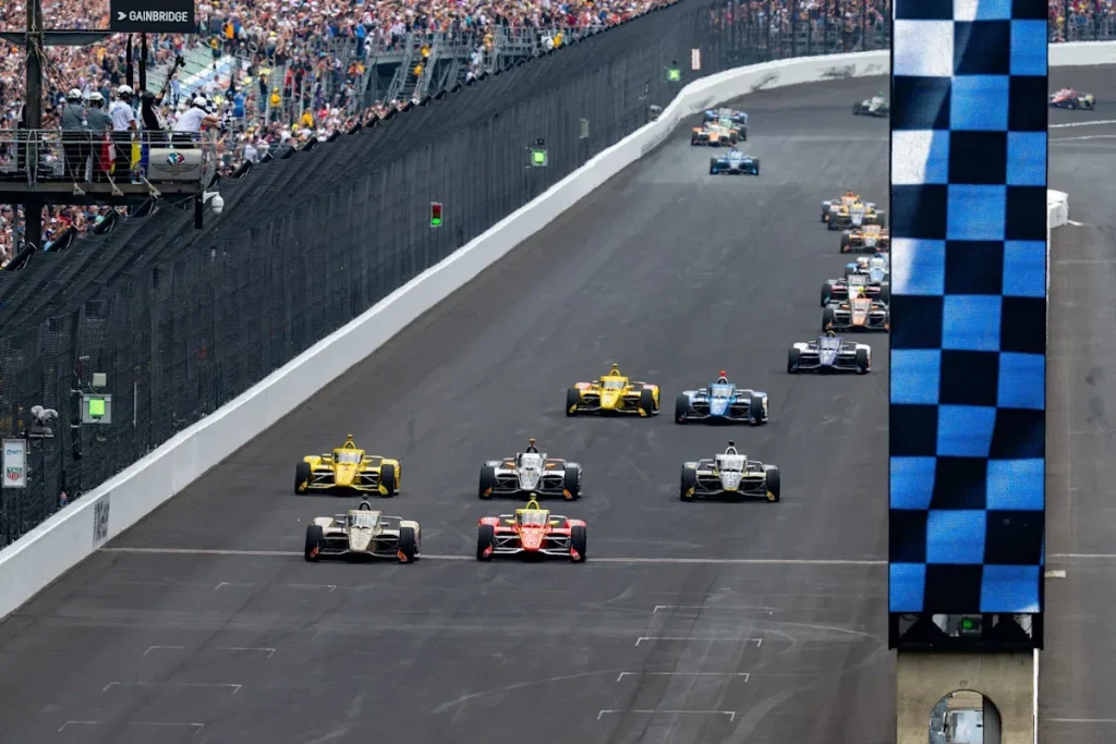 Open-wheel racing cars spread across the Indianapolis main straight beside crowded grandstands and a flag stand.