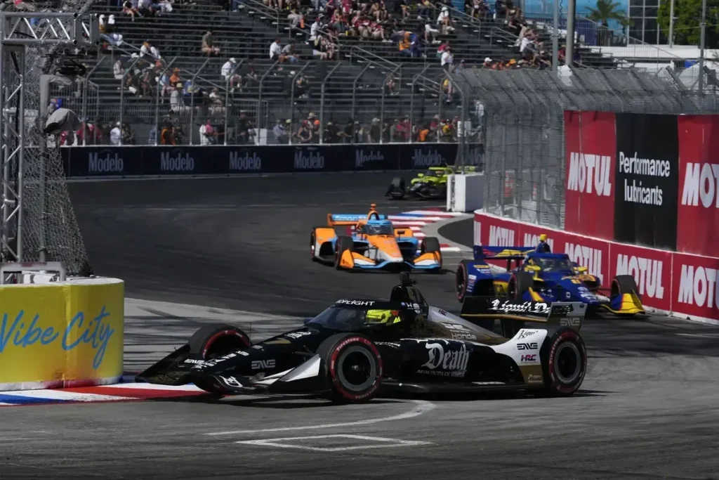 A black-and-white open-wheel racing car rounds a street-circuit corner, with blue-and-yellow and orange cars behind it beside barriers and catch fencing.