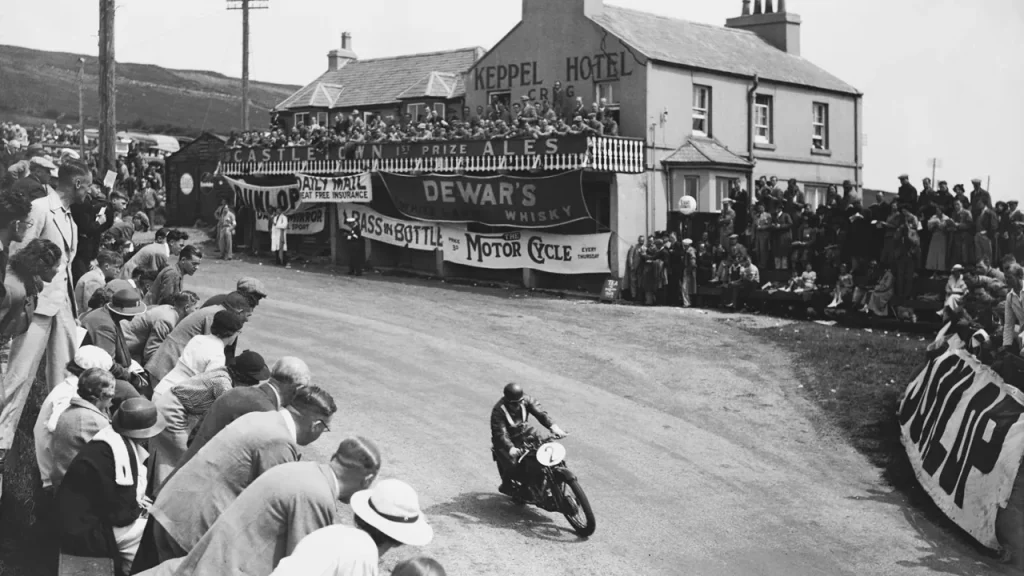 A motorcycle racer on a vintage motorcycle leans into a turn on a road lined with spectators outside a hotel building.