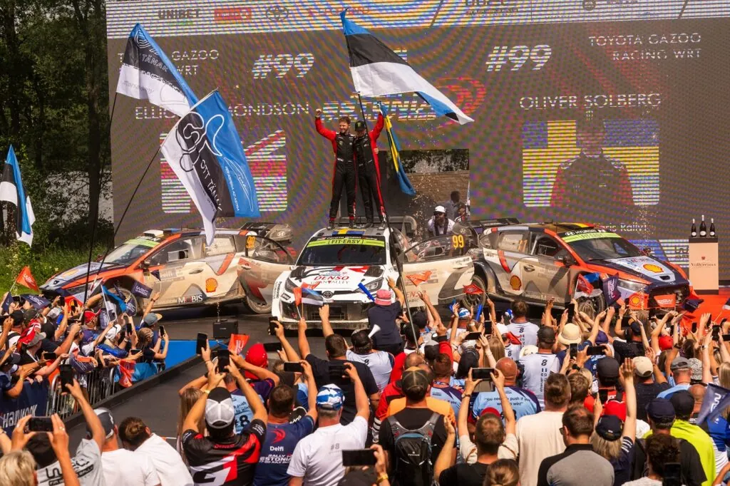 Rally drivers celebrating atop a rally car on a podium stage in front of a crowd holding flags and cameras.