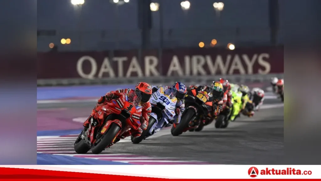 A line of motorcycle racers leaning through a turn on an illuminated asphalt track at night past a trackside barrier.