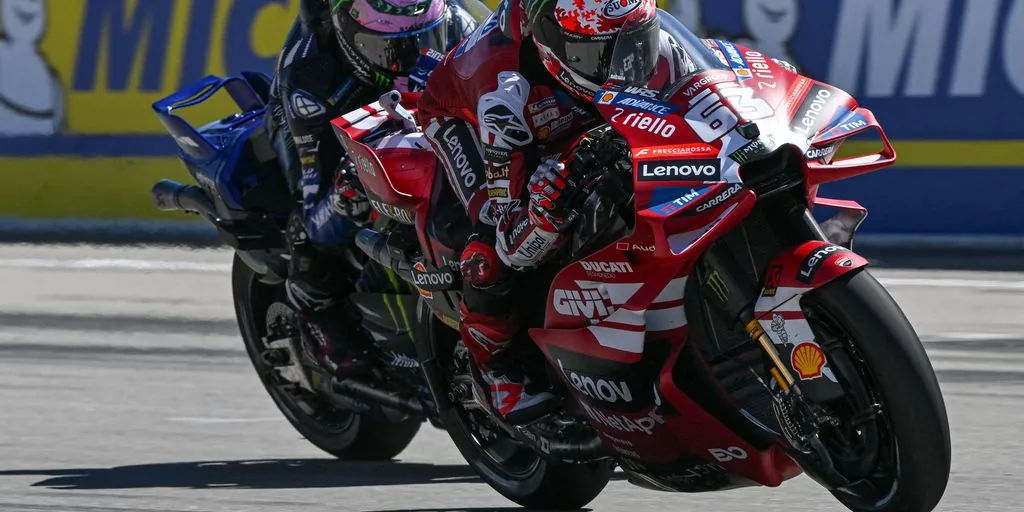 A red racing motorcycle equipped with front aerodynamic winglets leans through a corner on a circuit closely followed by another competitor.