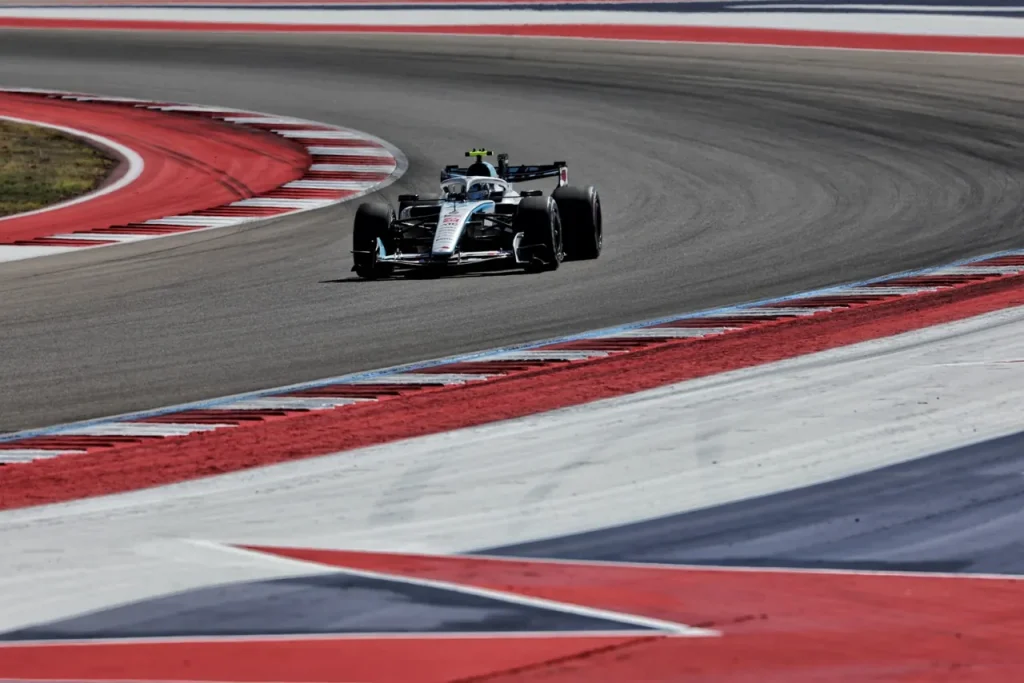 An open-wheel formula racing car navigates a turn on an asphalt racetrack featuring red-and-white kerbing and red, white, and blue painted run-off areas.