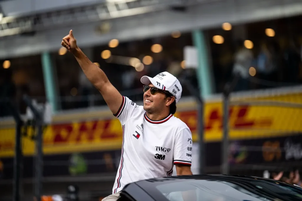 A smiling man wearing sunglasses, a white Cadillac cap, and a white team shirt giving a thumbs-up from an open-top car.