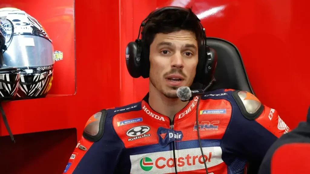 A motorcycle racer wearing a red, blue, and white racing suit and a headset with a microphone seated in a garage.