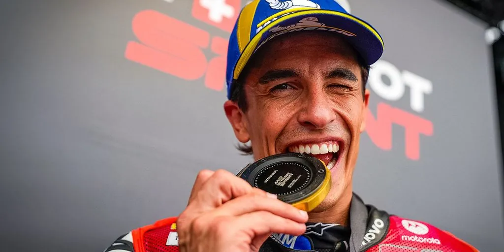 A motorcycle racer smiles and bites a sprint winner medal while celebrating.