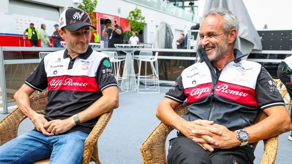 Two men wearing Alfa Romeo team apparel seated outdoors in wicker chairs and smiling.