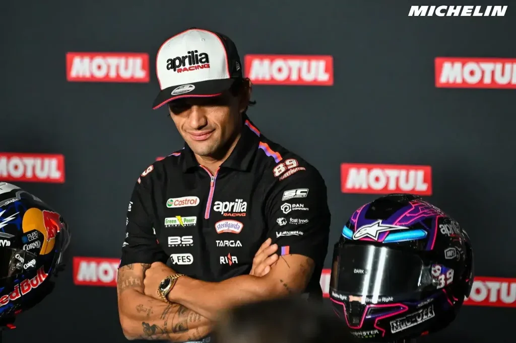 A motorcycle racer wearing an Aprilia Racing cap and team shirt stands with crossed arms beside a racing helmet in front of a sponsor backdrop.