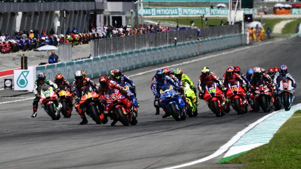 A pack of motorcycle racers riding down an asphalt track alongside a pit wall and grandstand.