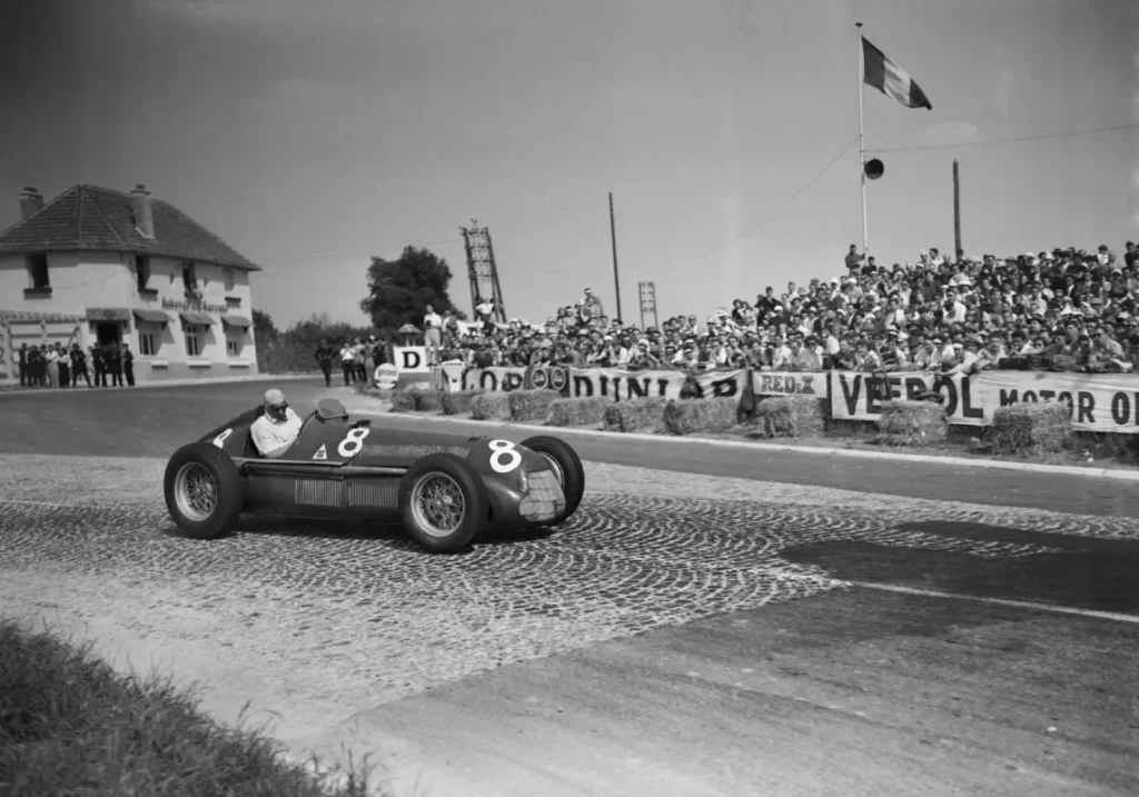 Black-and-white photograph of an open-wheel number 8 racing car on a cobbled corner, with spectators behind hay bales.