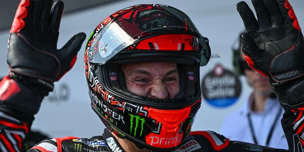 Close-up of MotoGP rider Fabio Quartararo wearing an HJC helmet with the visor raised and Monster Energy Yamaha racing gear, celebrating with both gloved hands raised in parc fermé or the pit lane.