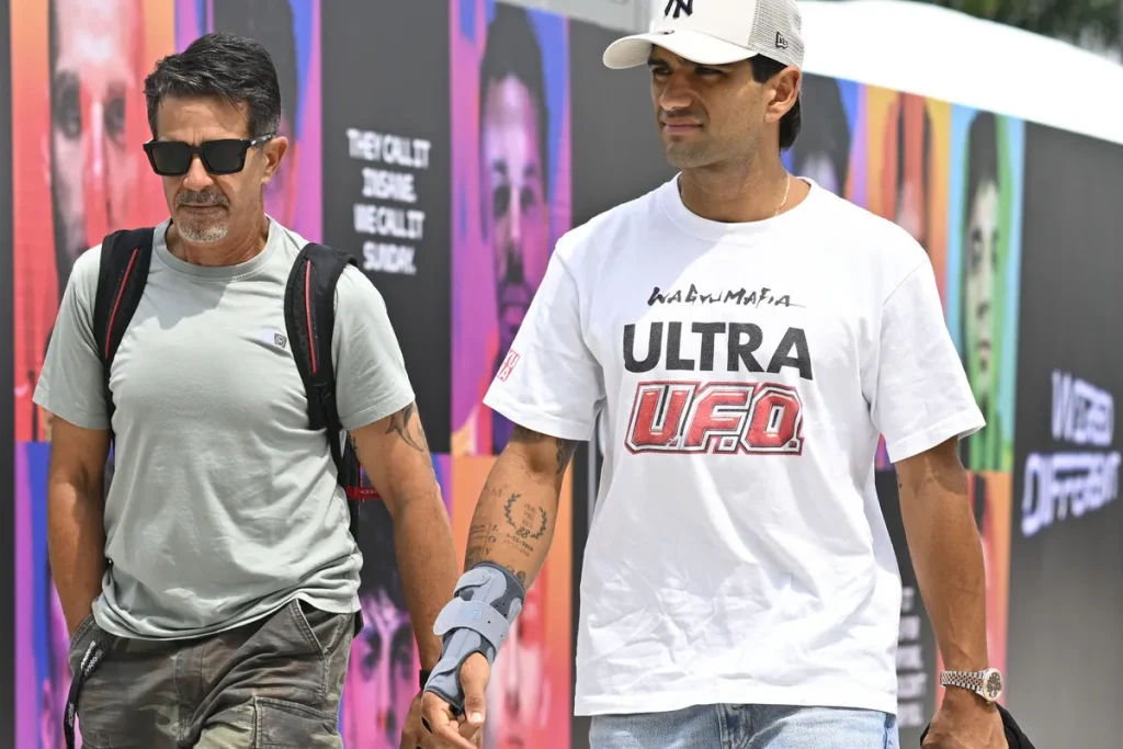 Man wearing a white t-shirt, baseball cap, and wrist brace walking beside a man wearing sunglasses.