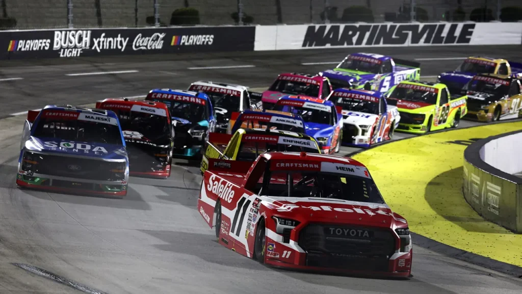 A pack of racing pickup trucks cornering on an asphalt track in front of an outer wall marked Martinsville.