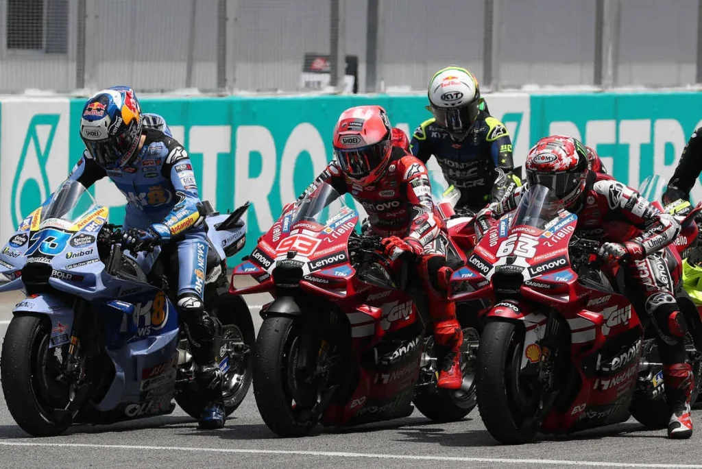 Motorcycle racers in protective gear seated on racing motorcycles lined up on an asphalt track in front of a barrier.