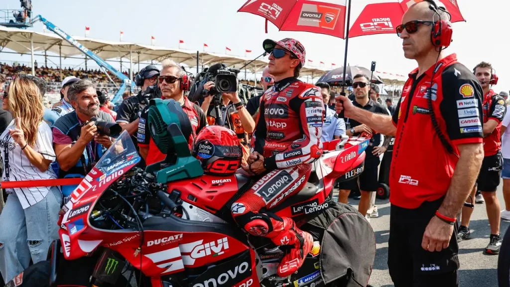 A motorcycle racer in a red racing suit and cap sits on a red racing motorcycle on a starting grid, surrounded by team crew members holding an umbrella and a cooling fan.
