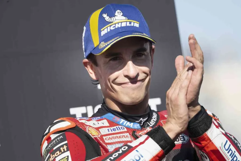 A smiling motorcycle racer wearing a blue Michelin cap and red racing leathers, clapping his hands.