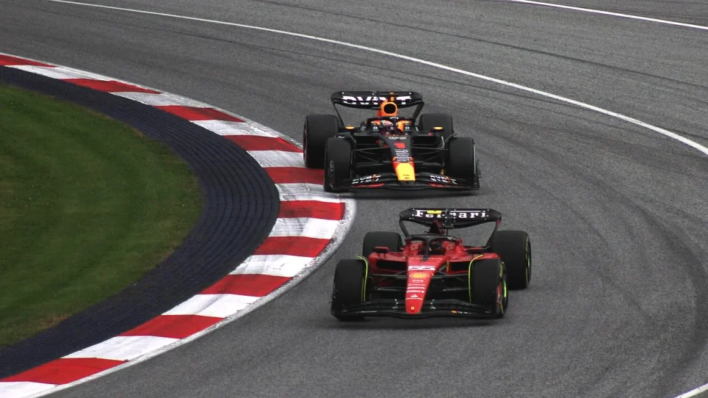 A red Ferrari leads a dark Red Bull through a circuit corner beside red-and-white kerbing and grass.