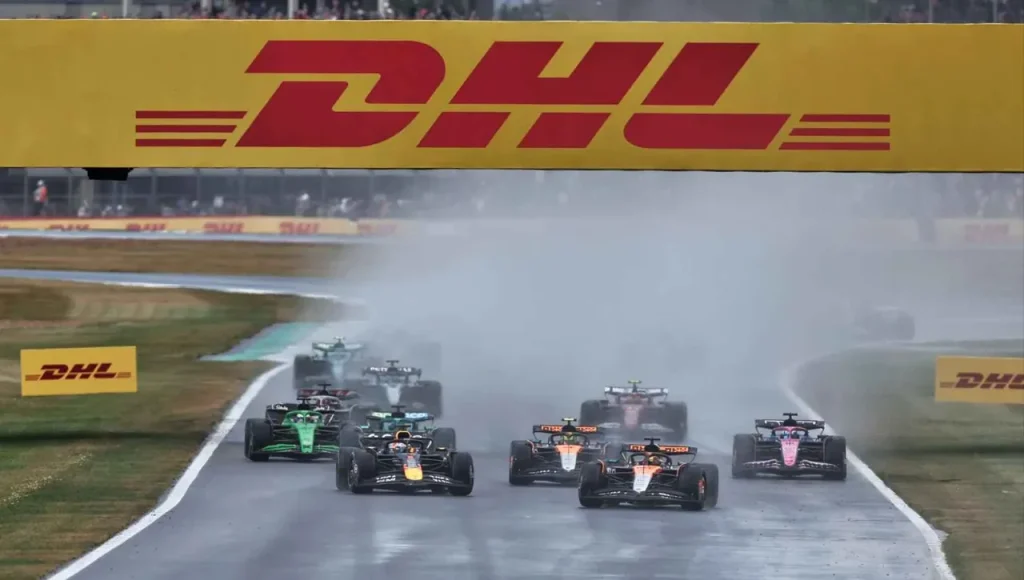 Formula 1 cars racing on a wet track under a yellow DHL overhead gantry.